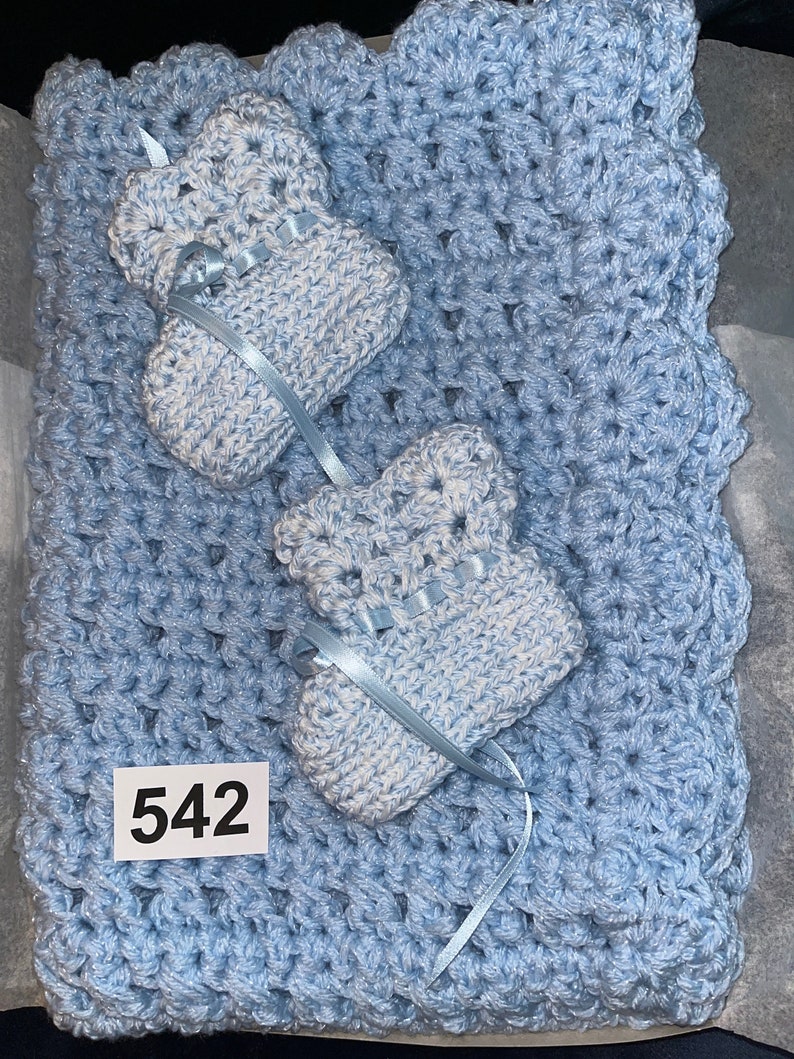May include: A light blue crocheted baby blanket with two matching booties. The blanket has a scalloped edge and the booties have a white and blue pattern. The number 542 is written on a white tag attached to the blanket.