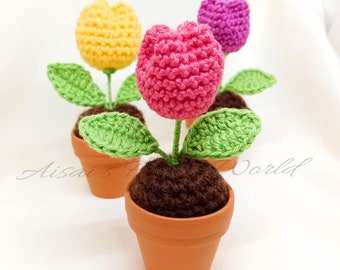 以下が含まれることがあります： テラコッタ製の鉢に植えられた、3つの手編みのチューリップの植物。チューリップはピンク、イエロー、パープルです。植物は白い表面の上に置かれています。