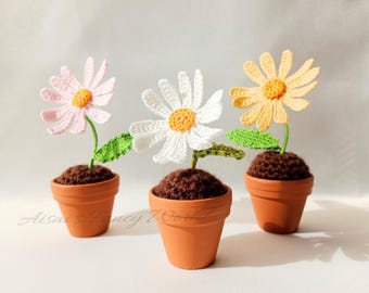 以下が含まれることがあります： 3つのポットに入れた、かぎ針編みのデイジーの花。デイジーは白、ピンク、黄色。ポットは茶色です。