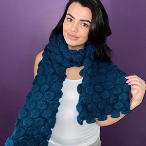 May include: A teal blue knitted scarf with a textured, bubble-like pattern. The scarf is long and appears to be made of a soft, warm material. The scarf is wrapped around the neck and shoulders.
