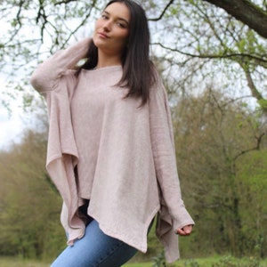 May include: A woman wearing a light pink, asymmetrical sweater with long sleeves and a loose fit. She is wearing blue jeans and is standing in a grassy area with trees in the background.