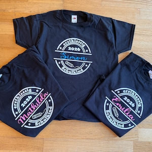 Könnte beinhalten: Drei schwarze T-Shirts mit einer kreisförmigen weißen Grafik, die den Text "erstklassig 2026 Schulkind" und ein Bleistift-Symbol enthält. Jedes Shirt zeigt einen individuellen Namen in einer anderen Farbe: "Simon" in Blau, "Mathilda" in Pink und "Emilia" in Pink.