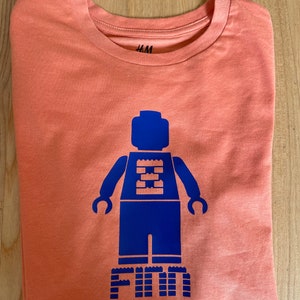 Könnte beinhalten: Ein korallenfarbenes T-Shirt mit einer blauen Lego-Figur und dem Namen "Finn", der in blauen Lego-Steinen geschrieben ist.