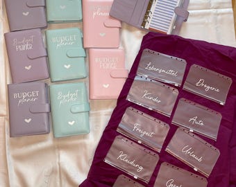 Könnte beinhalten: Ein Set aus neun pastellfarbenen Budgetplanern mit dem Aufdruck "Budget Planner" auf der Vorderseite. Die Planer sind in drei Reihen zu je drei angeordnet. Es gibt auch durchsichtige Plastikfächer mit Beschriftungen für verschiedene Ausgabenkategorien, wie z. B. "Lebensmittel", "Drogerie", "Kinder", "Freizeit", "Kleidung", "Auto", "Urlaub", "Geschenke" und "Einkauf".