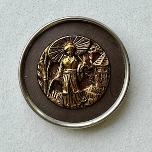 May include: A gold-toned metal button with a raised design of a woman in a traditional Japanese dress holding an umbrella. The button is set against a brown background and has a silver-toned rim.