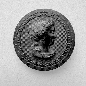 May include: A black, circular button with a detailed profile of a woman's face. The button has a textured background and a decorative border with geometric and circular patterns. The woman's hair is styled with a wreath.