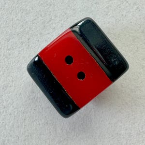May include: A square button with a red center stripe and black borders. The button has two small holes in the red section for sewing. The button is likely made of plastic or a similar material.