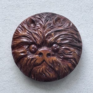 May include: A round, brown, wooden carving of a furry creature's face. The carving features detailed lines depicting fur, with visible eyes, nose, and ears. The overall design is circular and the wood has a natural, warm tone.