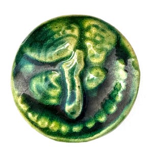 May include: A round, ceramic face with a smiling expression. The face is primarily green with yellow and dark green accents, and a glossy finish. The design features a nose, eyes, and a mouth with teeth.