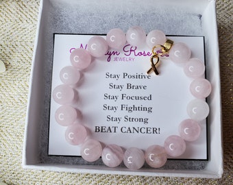 May include: A pink rose quartz bracelet with a gold charm in the shape of a ribbon. The bracelet is in a white box with a card that reads "Stay Positive, Stay Brave, Stay Focused, Stay Fighting, Stay Strong, BEAT CANCER!"