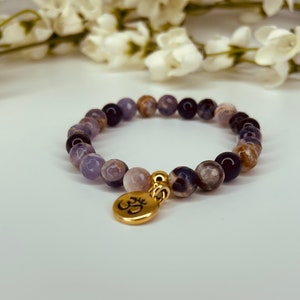 May include: A purple and blue gemstone bracelet with a gold Om charm. The bracelet is made of round beads and is strung on a stretchy cord.