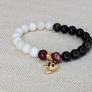 May include: A white and black beaded bracelet with a gold teapot charm. The beads are arranged in a yin yang pattern with white on one side and black on the other. The teapot charm is attached to the bracelet with a red bead.