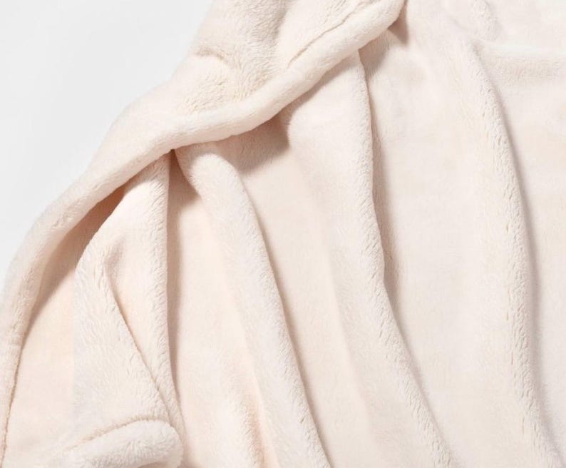 May include: A soft, white plush blanket with a smooth, fuzzy texture. The blanket is folded and draped over a surface, showcasing its luxurious feel.
