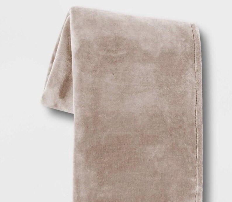 May include: A soft, plush throw blanket in a light beige color. The blanket is folded and draped over a white surface.