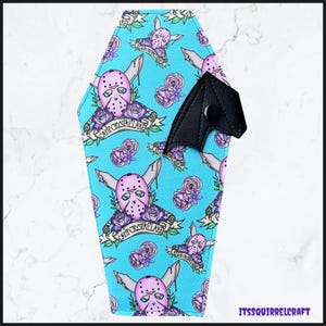 May include: A coffin-shaped accessory with a teal background featuring a repeating pattern of a pink hockey mask, knives, and purple roses. The text "CAMP CRYSTAL LAKE" is on a banner. A black bat-wing shaped closure is on the right.