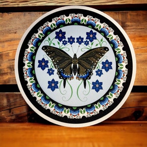 Puede incluir: Un plato redondo de cerámica blanco y negro con un diseño floral azul y verde y una mariposa negra cola de golondrina en el centro.