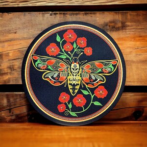 May include: A round, dark blue decorative item featuring a detailed moth design with red, yellow, and green accents. The moth is surrounded by red flowers and green vines, with a decorative border.