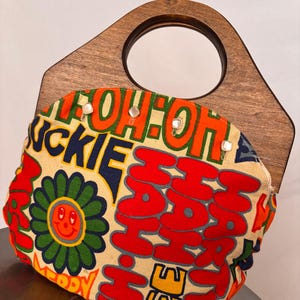 Puede incluir: Un bolso colorido con asa de madera. El bolso presenta una tela color crema con una flor y texto en rojo, naranja, amarillo y azul. El asa de madera tiene una abertura circular.