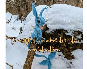 May include: A crochet pattern for a blue amigurumi creature with long ears and a tail. The creature is sitting on a branch in a snowy forest. The text "This is NOT a finished item: it's a crochet pattern" is visible in the image.