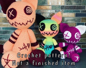 May include: Three crocheted plush toys in the shape of cats with stitched-on eyes, mouths, and X marks. The cat on the left is peach, the middle cat is green and yellow, and the cat on the right is purple. The text "Crochet pattern! Not a finished item" is at the bottom of the image.