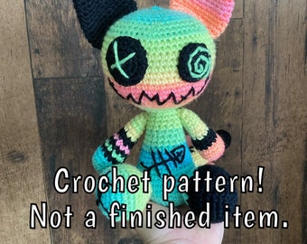 Può includere: Uno schema all'uncinetto per un giocattolo amigurumi di gatto colorato, verde, rosa e nero. Il gatto ha un viso cucito con occhi a forma di X e un design a spirale su un occhio. Il testo "Crochet pattern! Not a finished item." è visibile nell'immagine.