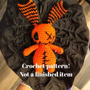 May include: Crochet pattern for a Halloween-themed orange and black amigurumi doll with stitched details and pins. "Crochet pattern! Not a finished item" text is visible.
