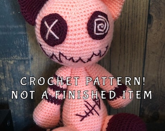 May include: Crochet pattern for a pink and burgundy cat amigurumi toy. The toy has stitched on eyes and a stitched on mouth. The text "CROCHET PATTERN! NOT A FINISHED ITEM" is visible in the image.