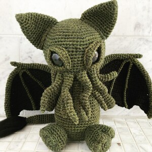 May include: Green crocheted plush toy with large eyes, a long tail, and bat-like wings.