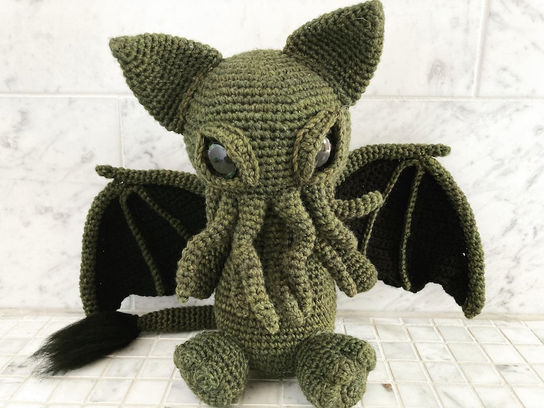 May include: Green crocheted plush toy with large eyes, a long tail, and bat-like wings.