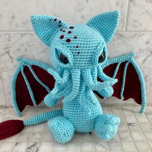 May include: A crocheted plush toy in the shape of a Cthulhu monster. The toy is light blue with maroon wings and a tail. It has large, black eyes and a tentacled head.
