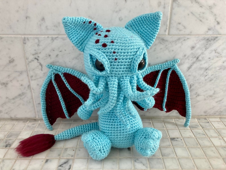 May include: A crocheted plush toy in the shape of a Cthulhu monster. The toy is light blue with maroon wings and a tail. It has large, black eyes and a tentacled head.