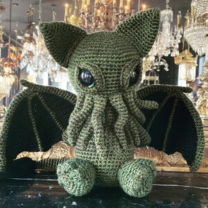 May include: Green crocheted plush toy with a cat-like face and large, black eyes. The toy has bat-like wings and a long, curled tail.