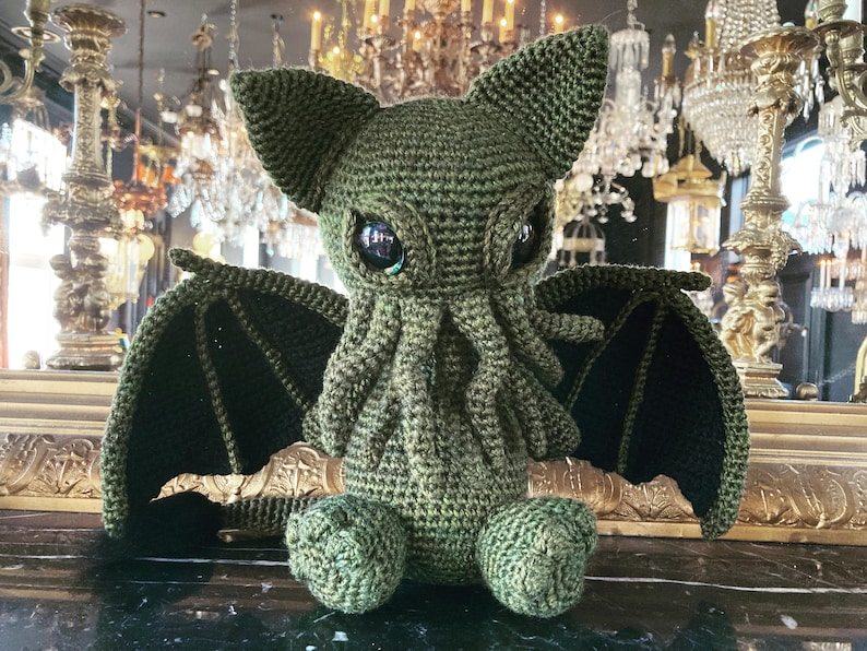 May include: Green crocheted plush toy with a cat-like face and large, black eyes. The toy has bat-like wings and a long, curled tail.