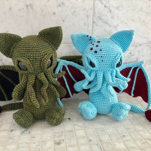 May include: Two crocheted plush toys shaped like Cthulhu, a fictional cosmic entity. One is green with black wings and a tail, the other is blue with maroon wings and a tail.
