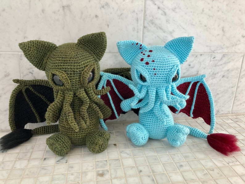 May include: Two crocheted plush toys shaped like Cthulhu, a fictional cosmic entity. One is green with black wings and a tail, the other is blue with maroon wings and a tail.