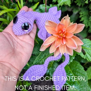 May include: A handmade, lavender crochet creature with large, black, glittery eyes. The creature is displayed with a peach-colored flower and green foliage. Text on the image reads: "THIS IS A CROCHET PATTERN, NOT A FINISHED ITEM."