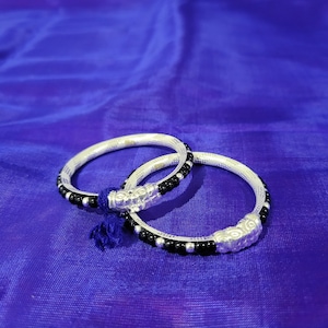 May include: Two silver bangles with black beads, one bangle is slightly larger than the other, both bangles have a decorative design on the front.