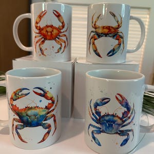 May include: Four white ceramic mugs with crab illustrations. Each mug features a different watercolor crab design in shades of orange, red, blue, and yellow. The mugs have a standard handle and are displayed on a white surface.