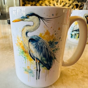 May include: White ceramic mug featuring a watercolor illustration of a great blue heron. The bird is depicted with blue, gray, yellow, and black feathers, with a long neck and beak. The mug has a curved handle and is suitable for hot beverages.
