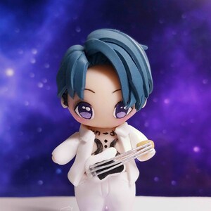 May include: A figurine of a cartoon character with blue hair, wearing a white suit and holding a white guitar. The figure stands on a purple star-shaped base. The background is a blurred purple and blue galaxy.