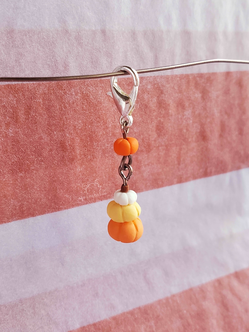 May include: A silver charm with a stack of three miniature pumpkins. The pumpkins are orange, yellow, and white. The charm is attached to a silver chain.