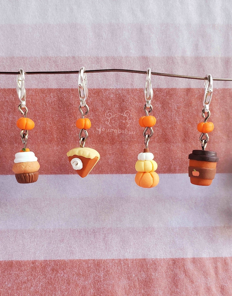 May include: Four fall-themed charms with orange and brown accents. The charms are attached to silver keychains and feature a cupcake, a pumpkin pie, a stack of pumpkins, and a coffee cup.