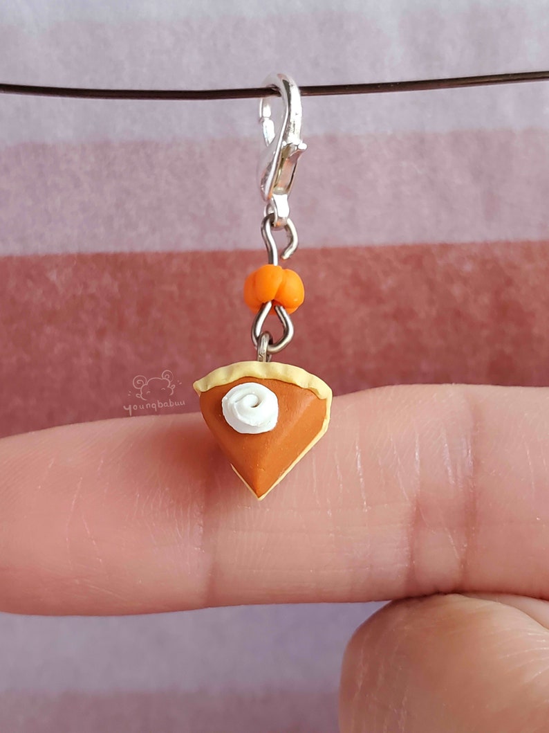 May include: A miniature pumpkin pie charm with a swirl of whipped cream on top. The charm is attached to a silver lobster clasp and a small orange bead.