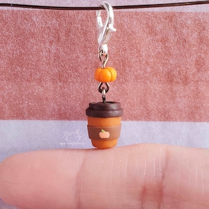 May include: A small, brown and orange coffee cup charm with a pumpkin design. The charm is attached to a silver lobster clasp.