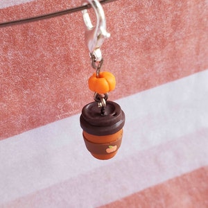 May include: A brown and orange polymer clay charm of a coffee cup with a pumpkin on top. The charm is attached to a silver lobster clasp.