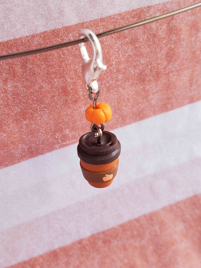 May include: A brown and orange polymer clay charm of a coffee cup with a pumpkin on top. The charm is attached to a silver lobster clasp.