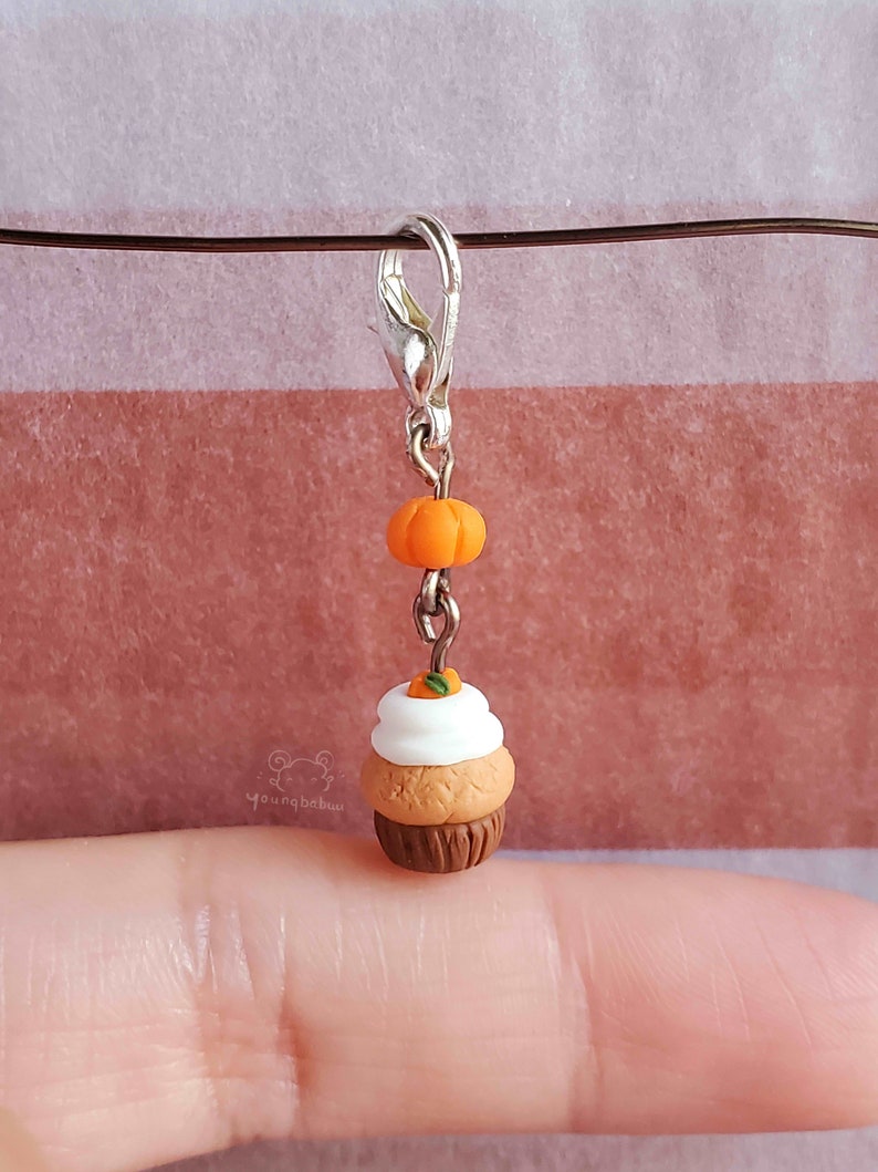 May include: A small, brown and white cupcake charm with a miniature orange pumpkin on top. The charm is attached to a silver lobster clasp.