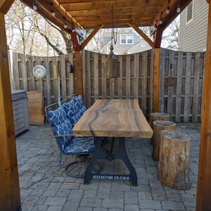 May include: A wooden table with a live edge top and black metal legs sits on a patio under a wooden pergola with string lights. The table is surrounded by three wooden stumps used as stools. The table has the text "JCS CASTING CO. CIN.O." printed on the bottom of the legs.