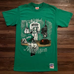 Puede incluir: Camiseta verde con un gráfico del equipamiento de fútbol de los Jets de Nueva York, que incluye una camiseta, un casco, un balón y botas. La camiseta tiene el texto "New York" y "Jets". La camiseta es de un material suave.
