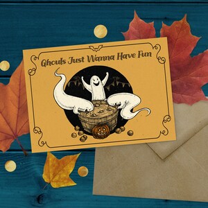 May include: A yellow card with the text "Ghouls Just Wanna Have Fun" and a cartoon illustration of three ghosts in a cauldron with a pumpkin. The card is on a brown envelope with a blue wooden background.